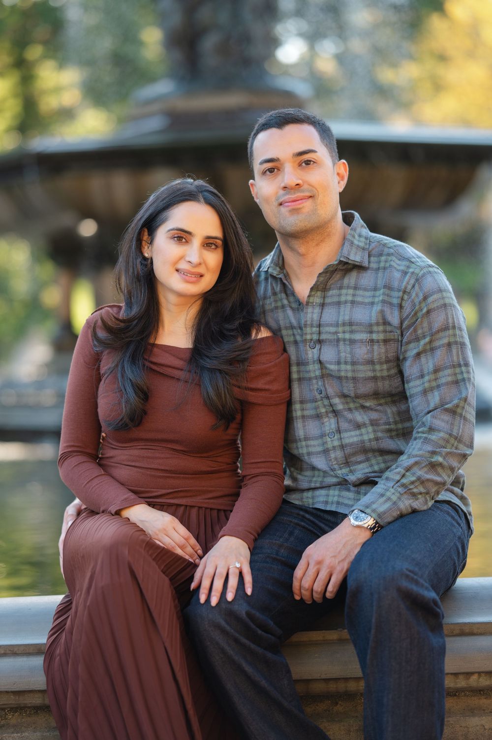 A couple sits together smiling in front of a decorative fountain, woman in brown dress, man in plaid shirt.