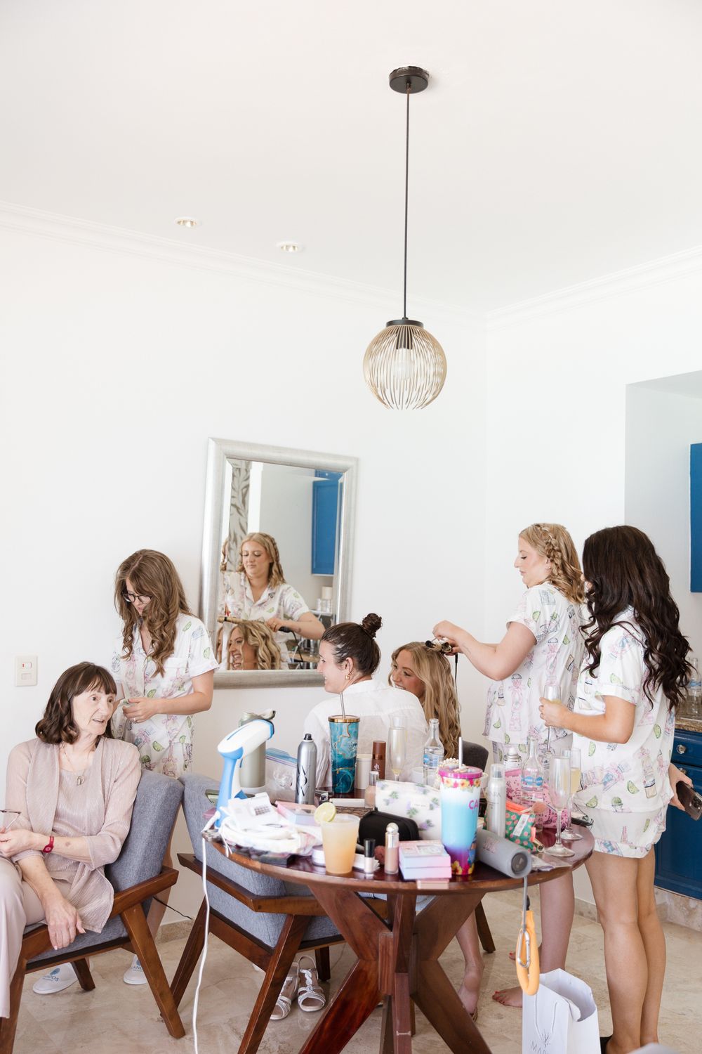 Bridesmaids in floral robes prepare for a wedding, styling hair around a table filled with beauty products in a bright white room.