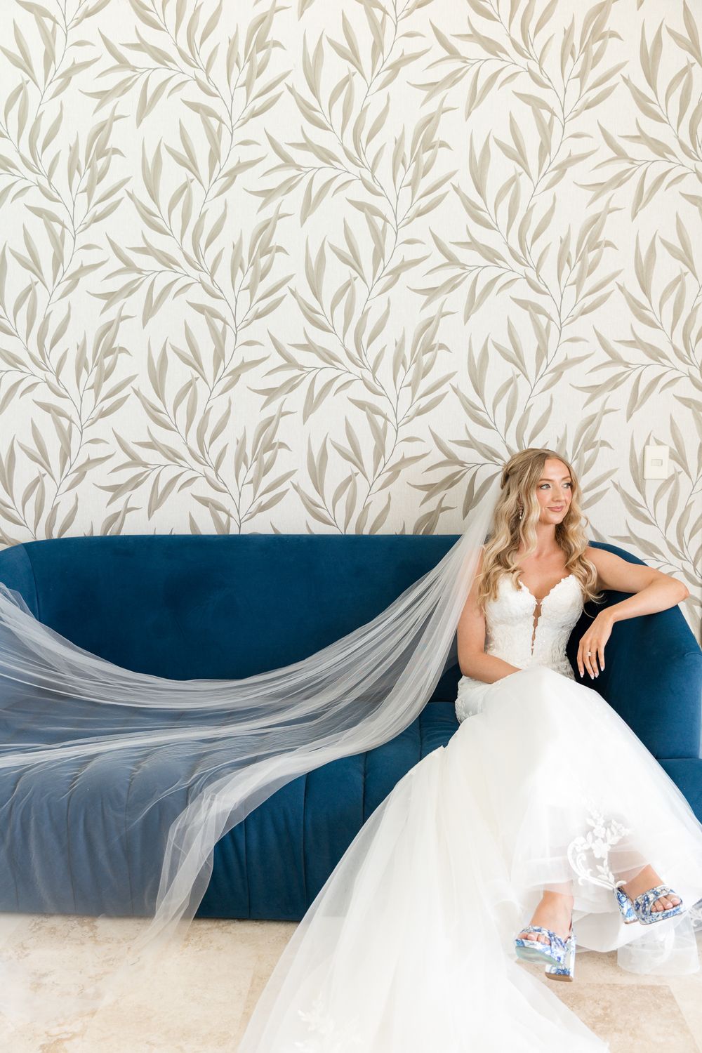 Bride in white ballgown and veil sits on blue velvet sofa against floral wallpaper wearing decorative heels.