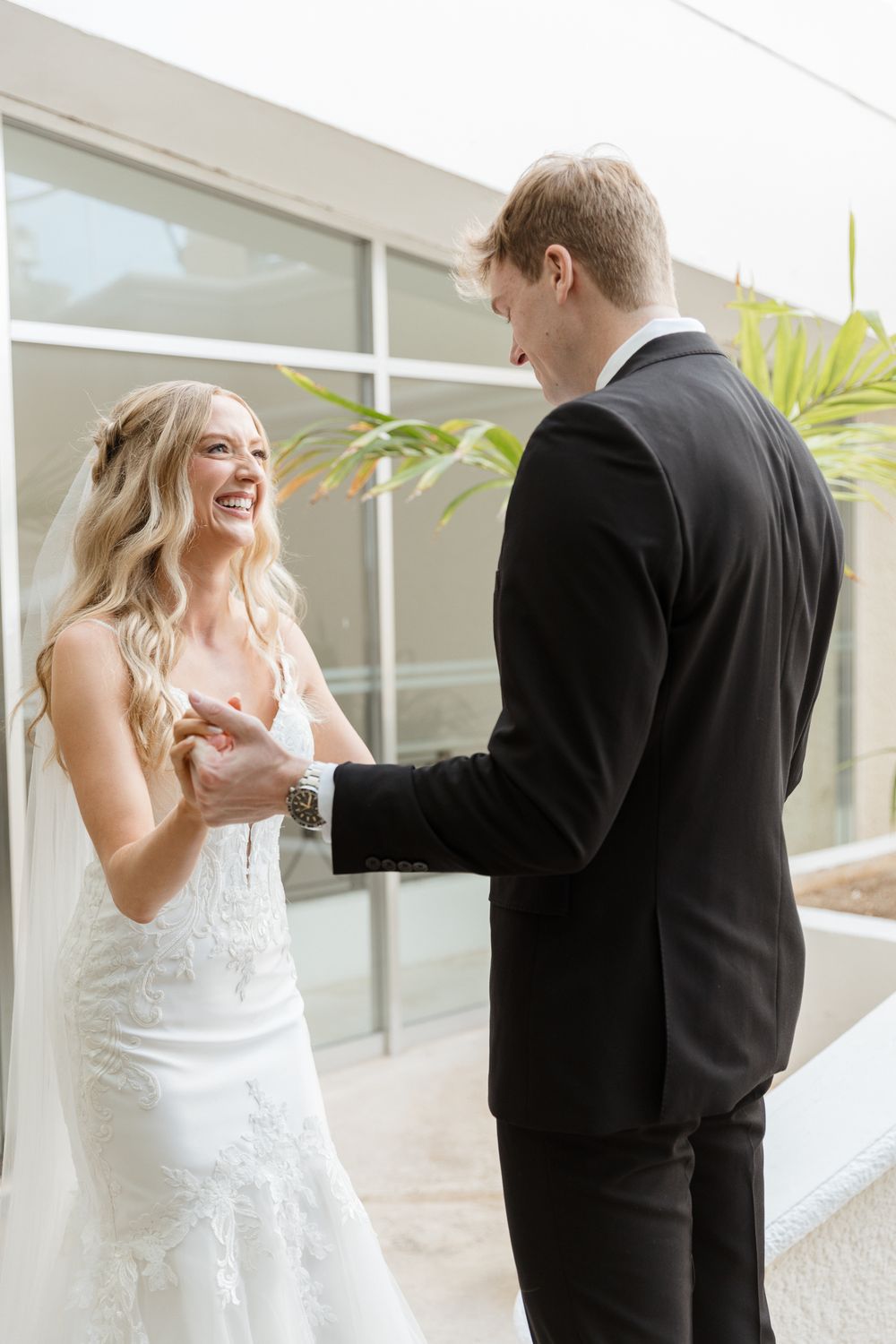 Bride in lace gown laughing joyfully during first look with groom in black tuxedo at wedding venue.