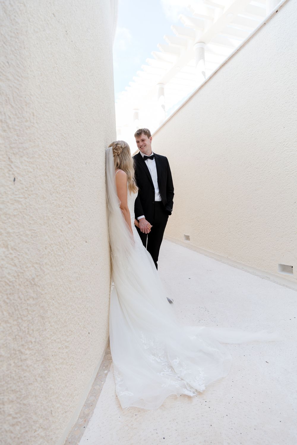 Bride and groom hold hands in narrow white alley, her long veil trailing on the ground.