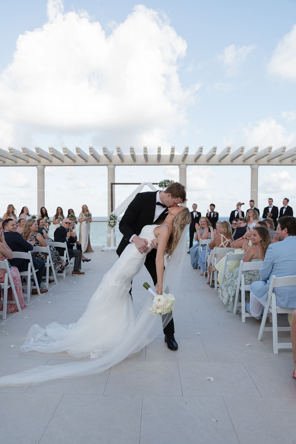 Bride and groom share a romantic dip kiss at outdoor rooftop wedding ceremony with guests seated on both sides.
