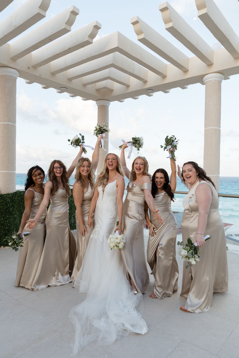 Bride with bridesmaids in champagne satin gowns celebrate under pergola with ocean view.