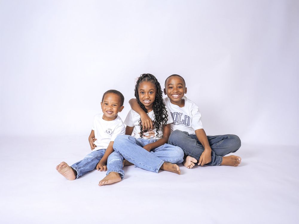 Three young children smiling and posing together in white shirts and jeans against a white studio background.