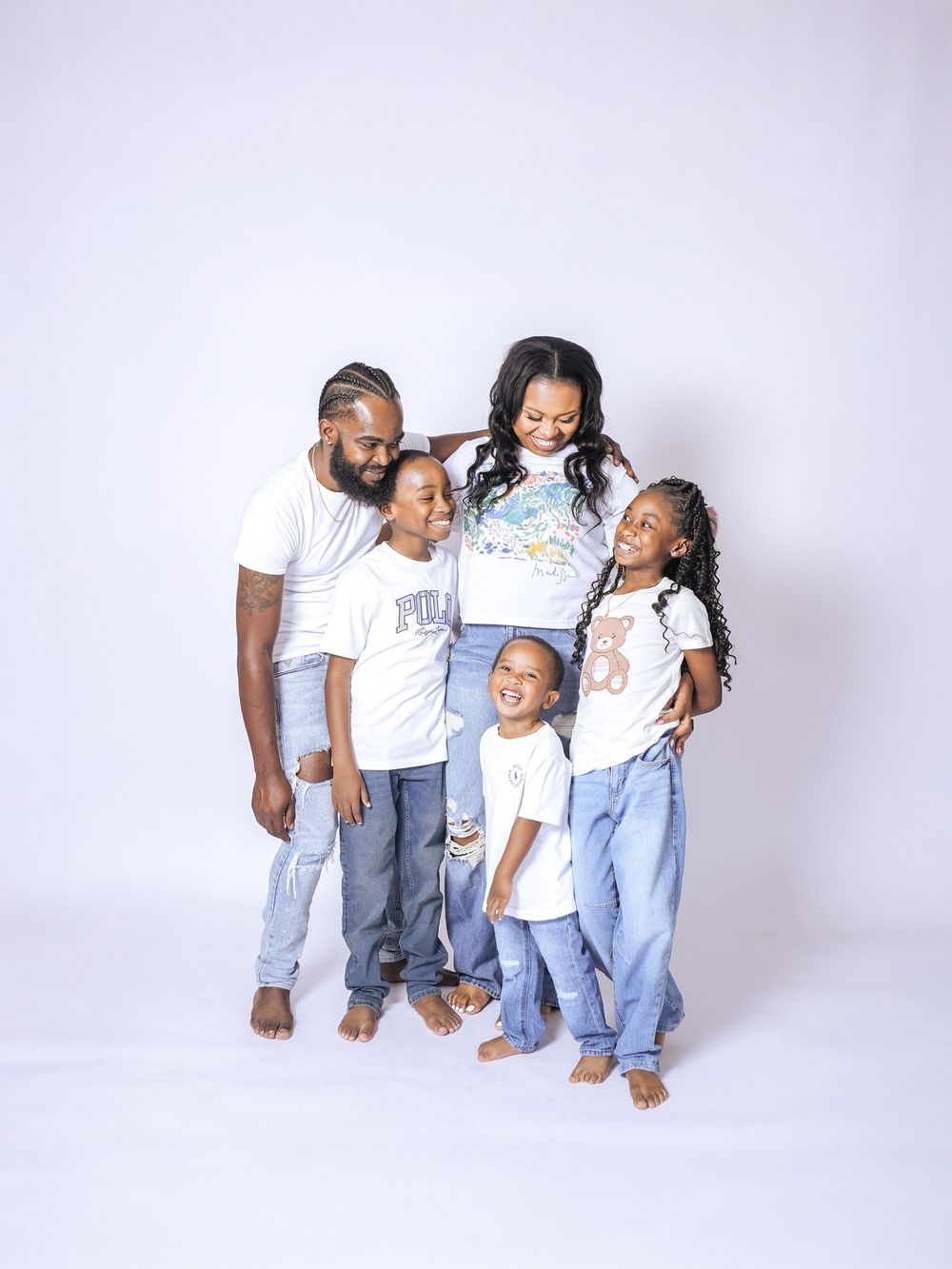 A joyful family of five in matching white tees and jeans poses together against a white studio background.