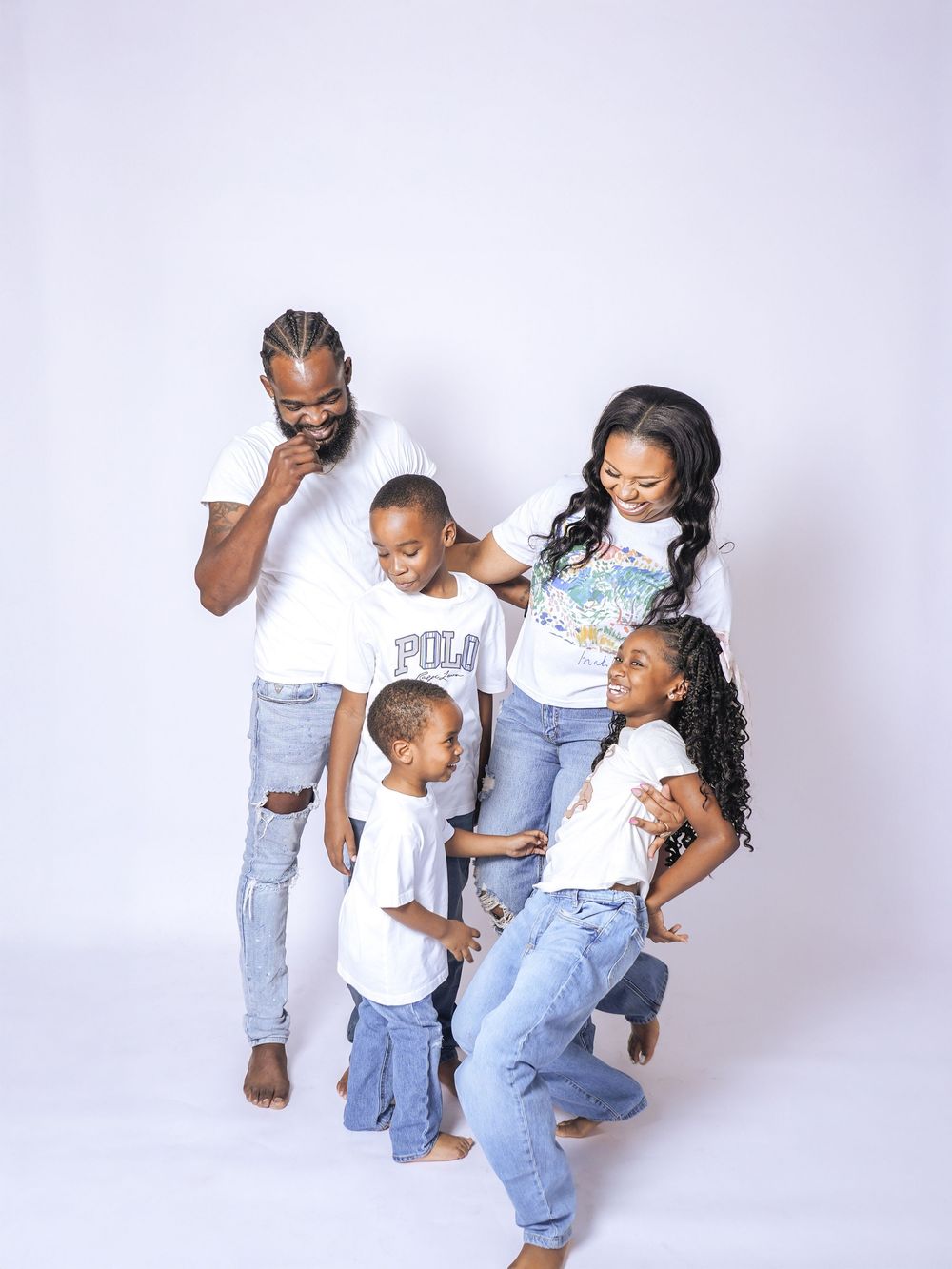 A joyful Black family of five in matching white tees and jeans poses playfully against a white studio backdrop.