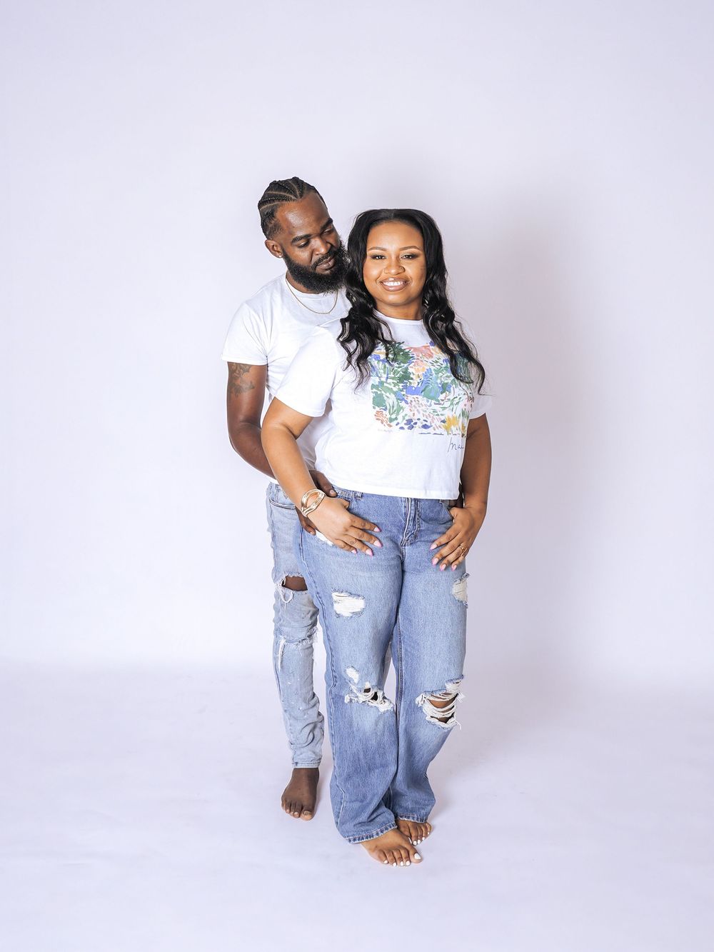 A couple poses together in matching white tees and ripped jeans against a white backdrop in a studio photo.