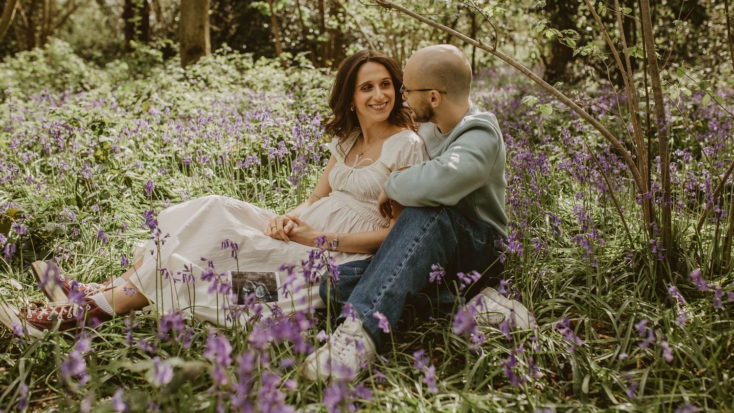 Couple sitting in a bluebell forest, woman in white dress with baby bump, man embracing her tenderly.