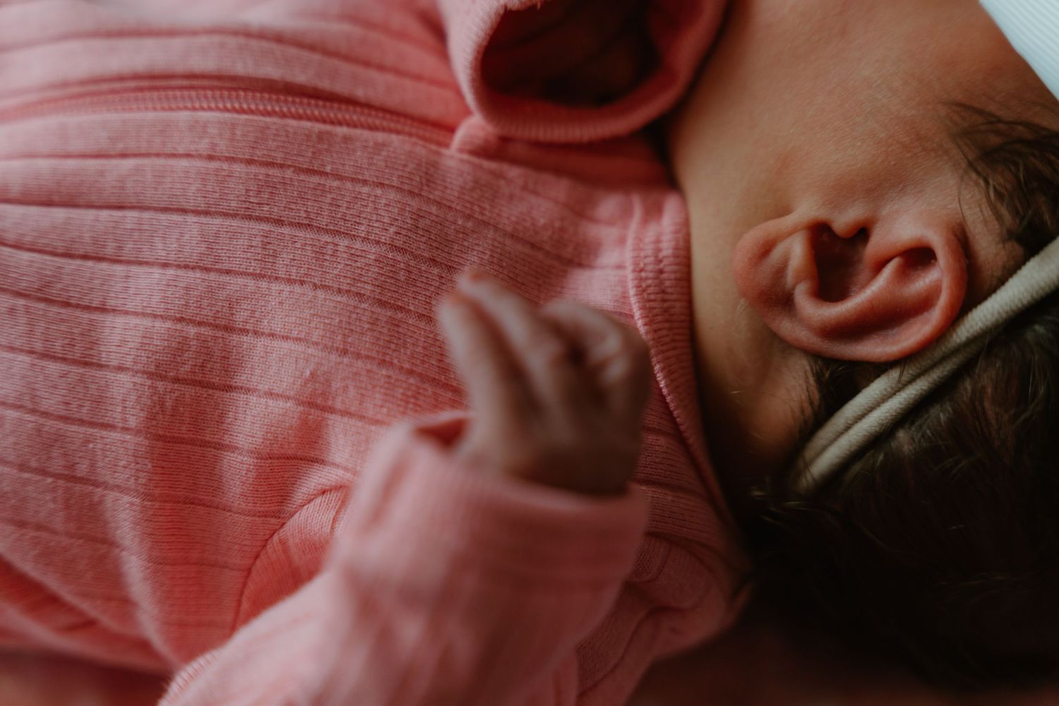 Close-up of sleeping newborn baby in pink ribbed outfit showing tiny hand and ear detail.