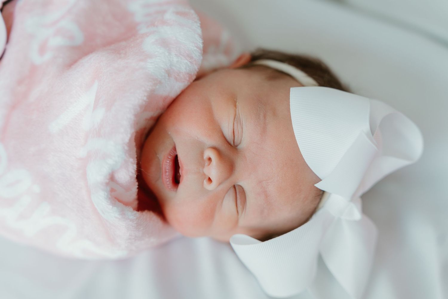 Peaceful newborn baby girl sleeping in pink blanket wearing white bow hospital hat.