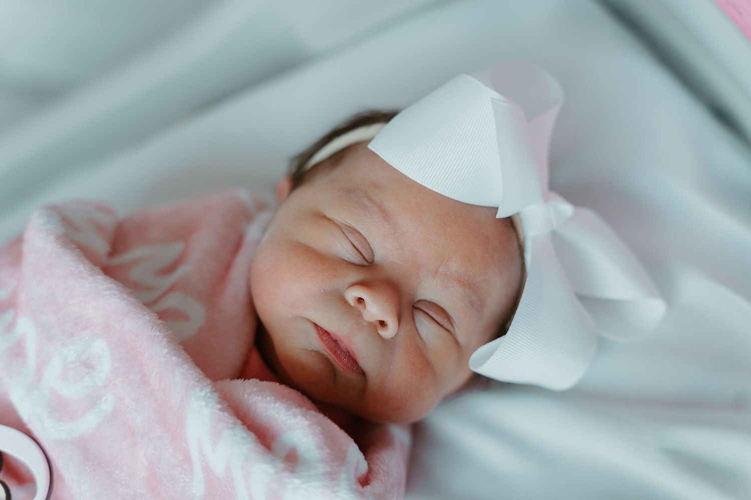 Serene newborn baby girl sleeping with white bow headband wrapped in soft pink blanket.
