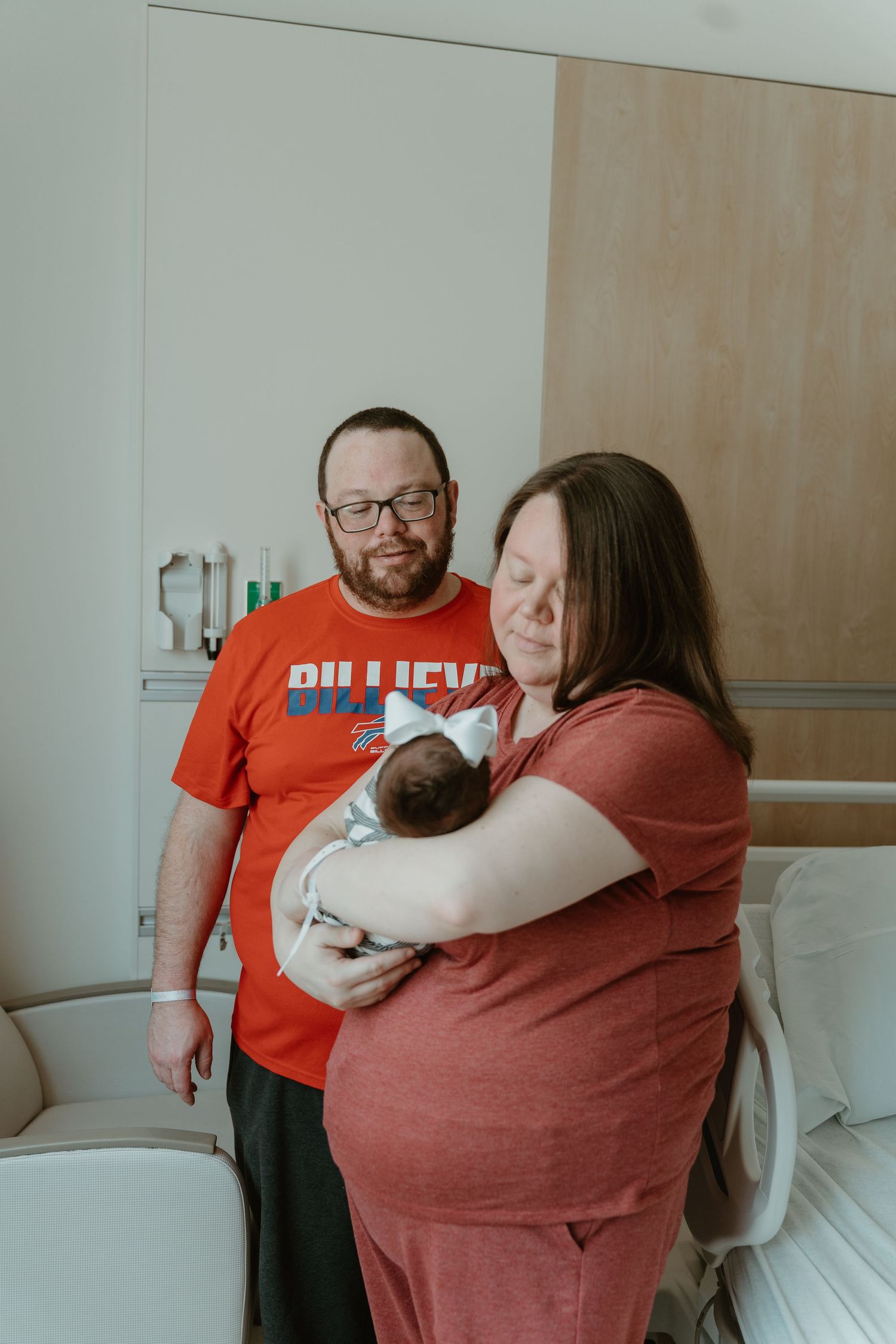 New parents in matching red shirts tenderly holding their newborn baby in hospital room.