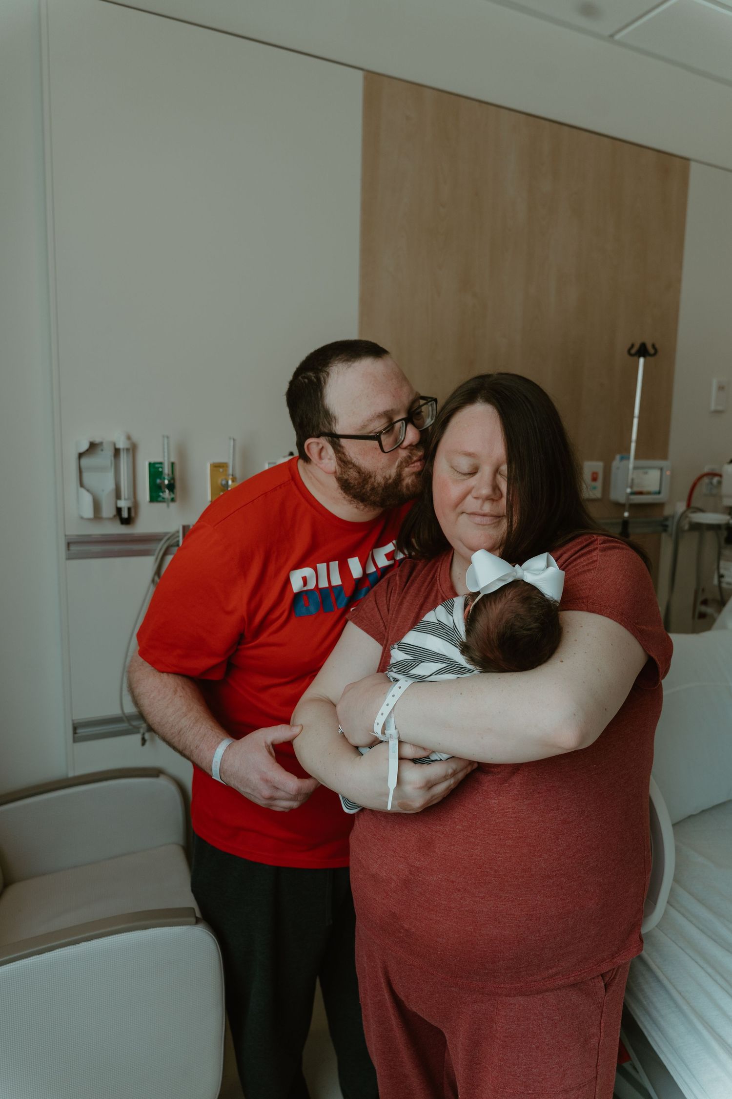 New parents share a tender moment in hospital room, father kissing mother's cheek as she cradles their newborn baby.