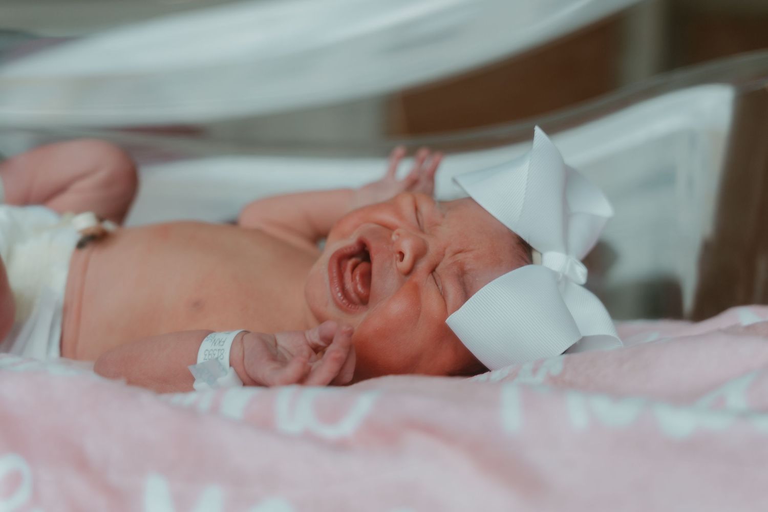 Crying newborn baby girl wearing white bow headband lying on pink blanket in hospital bassinet.