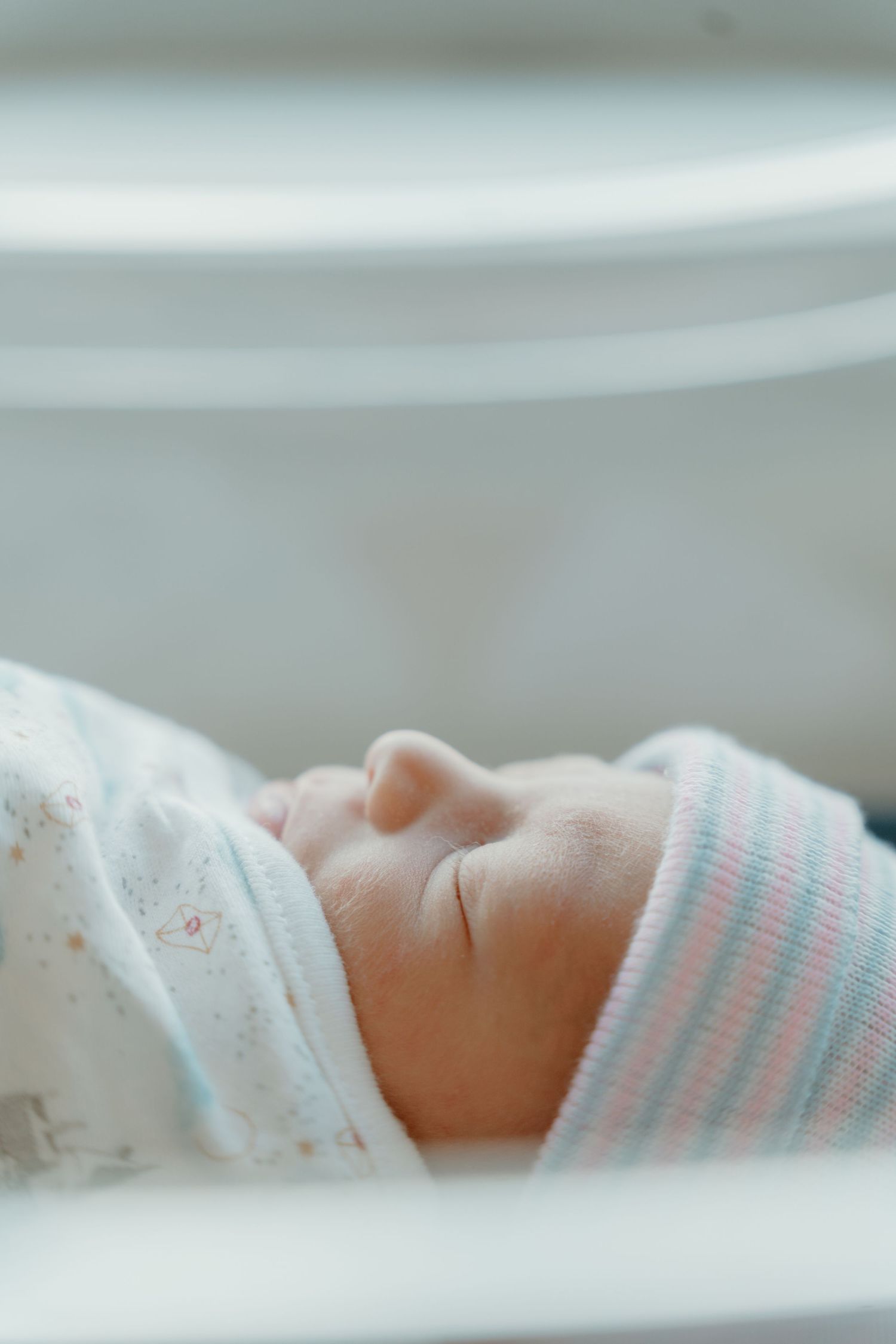 Newborn baby sleeping in a striped hospital hat, photographed through white crib rails.