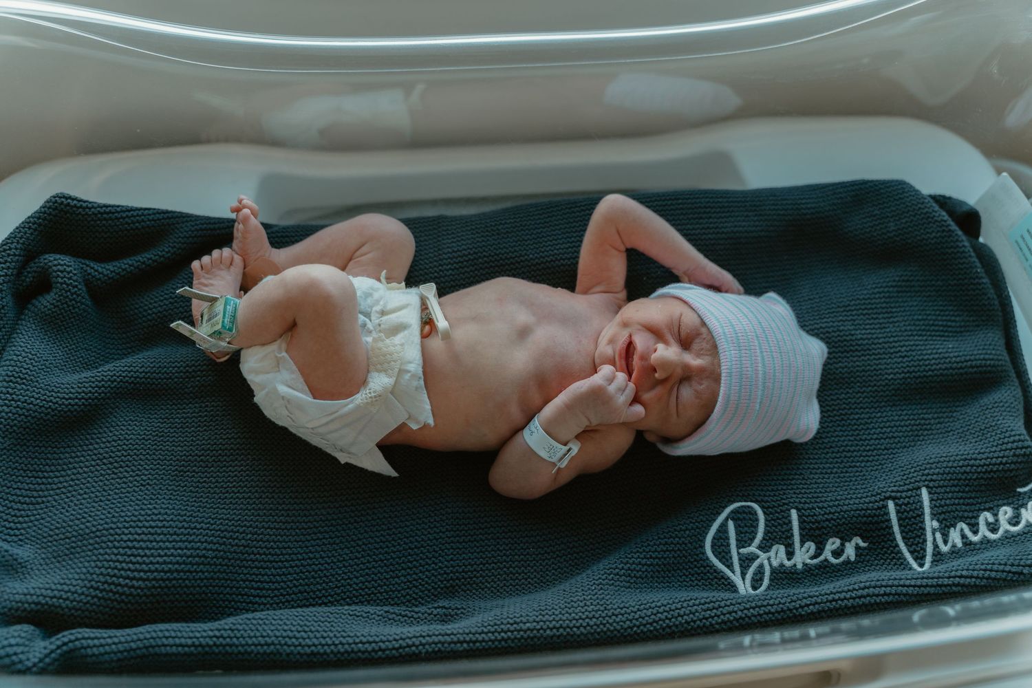 Newborn baby resting on a dark blanket embroidered with Baker Vincent in a hospital bassinet.