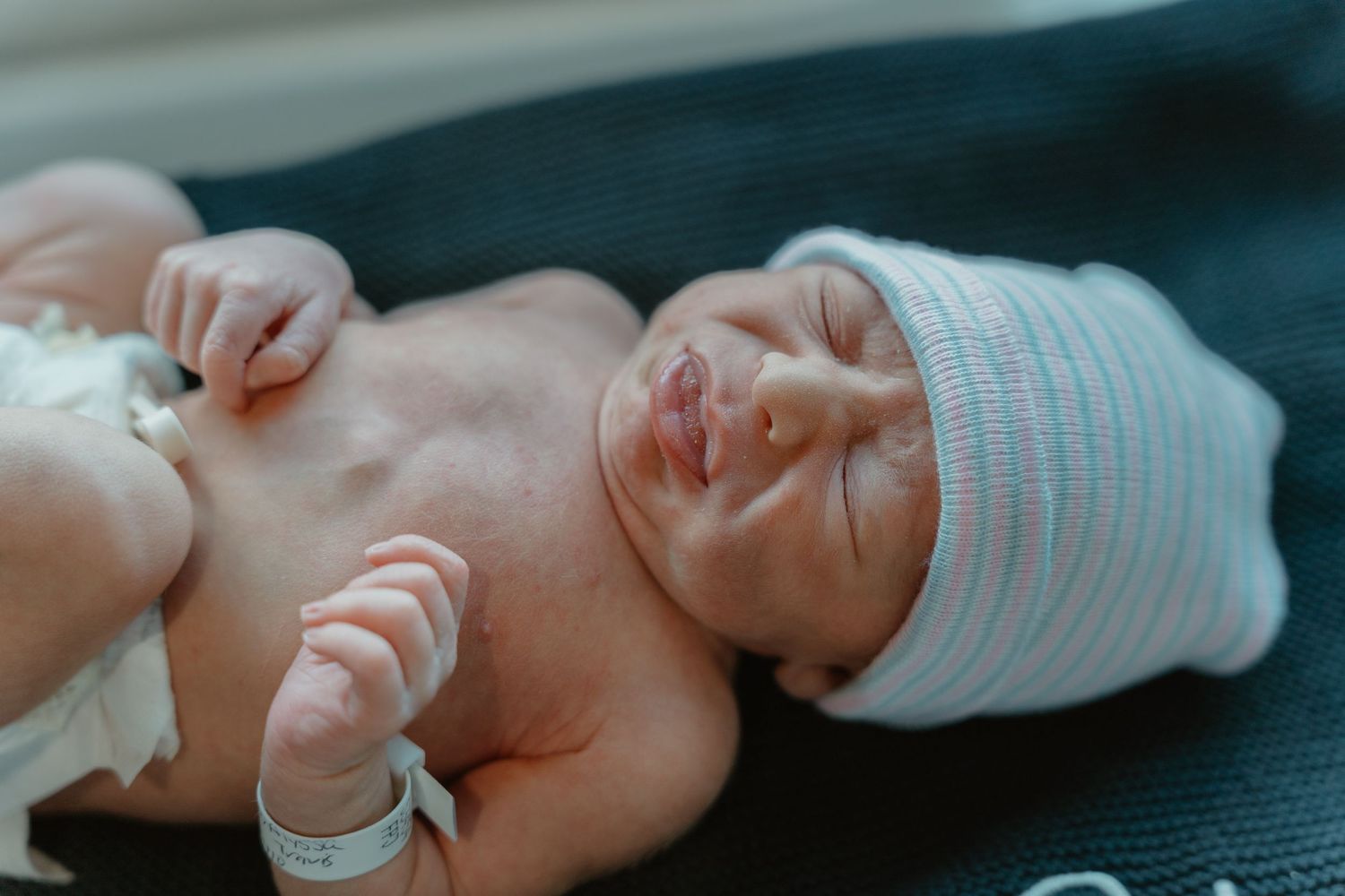 Peaceful newborn baby wearing a striped hospital hat lying on a dark blanket.