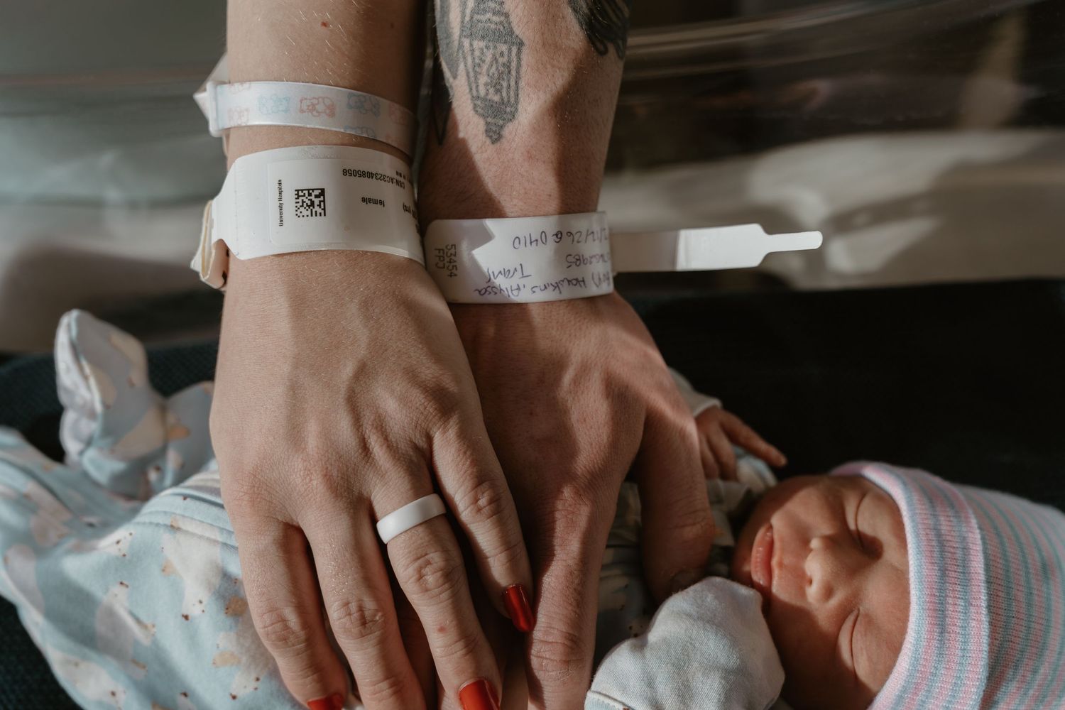 Close-up of tattooed mother's hands with hospital bracelets gently touching her newborn baby.