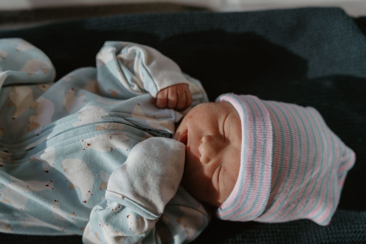 Sleeping newborn baby wearing a striped hat dressed in a light blue spotted onesie.