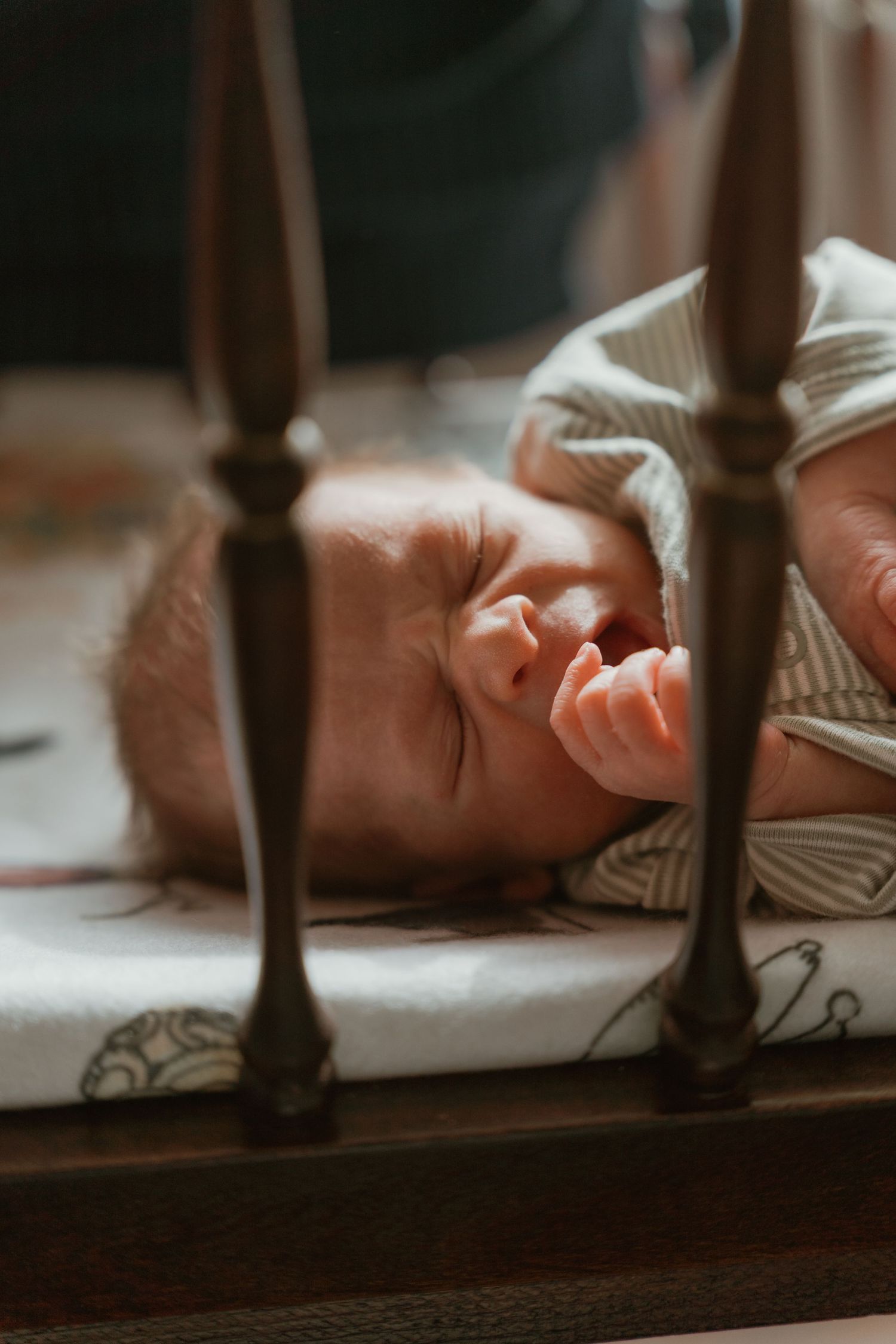 Newborn baby sleeping in wooden crib, photographed through crib rails on patterned mattress.