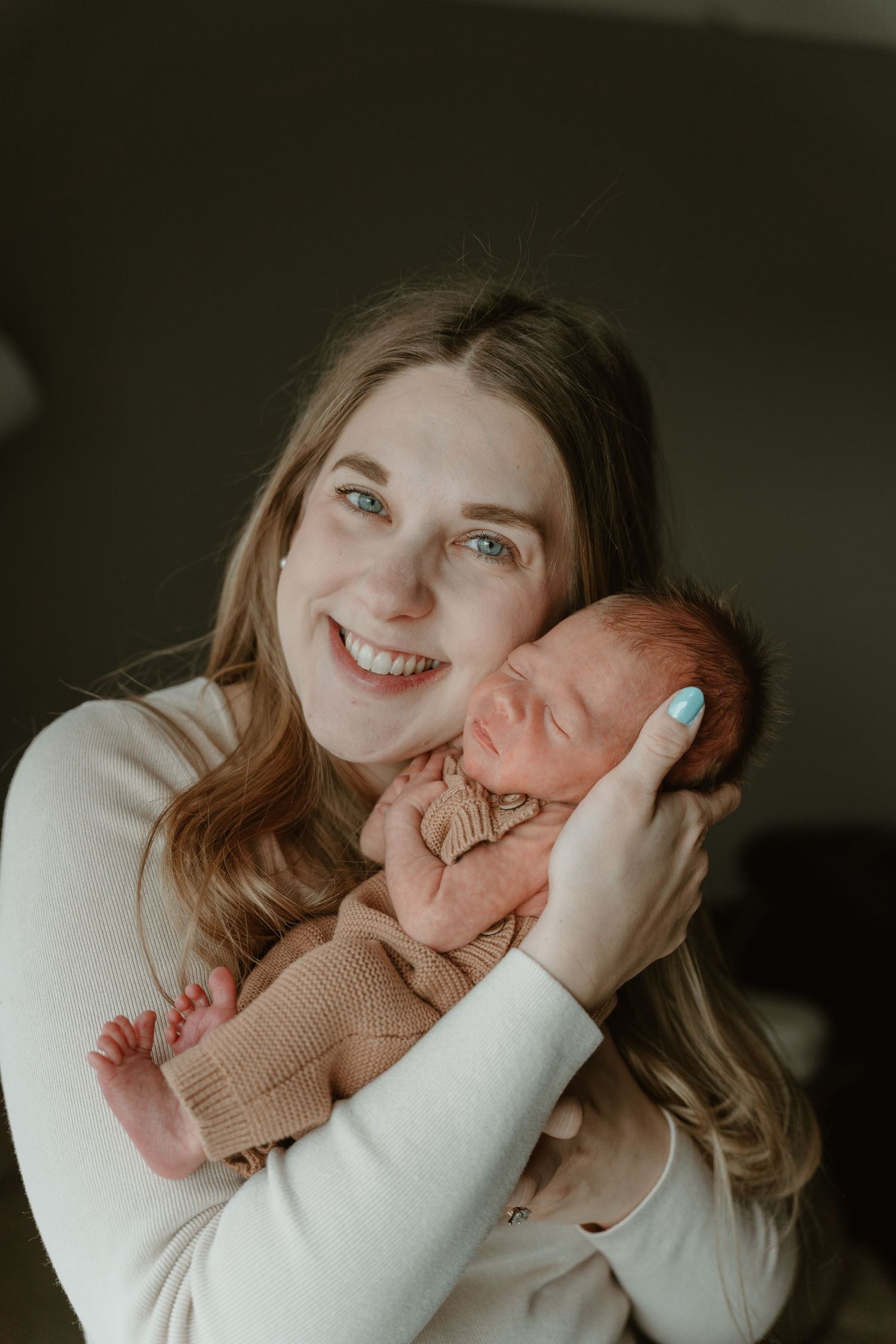 Smiling new mother with blue nails holding sleeping newborn baby dressed in tan outfit close to her cheek.