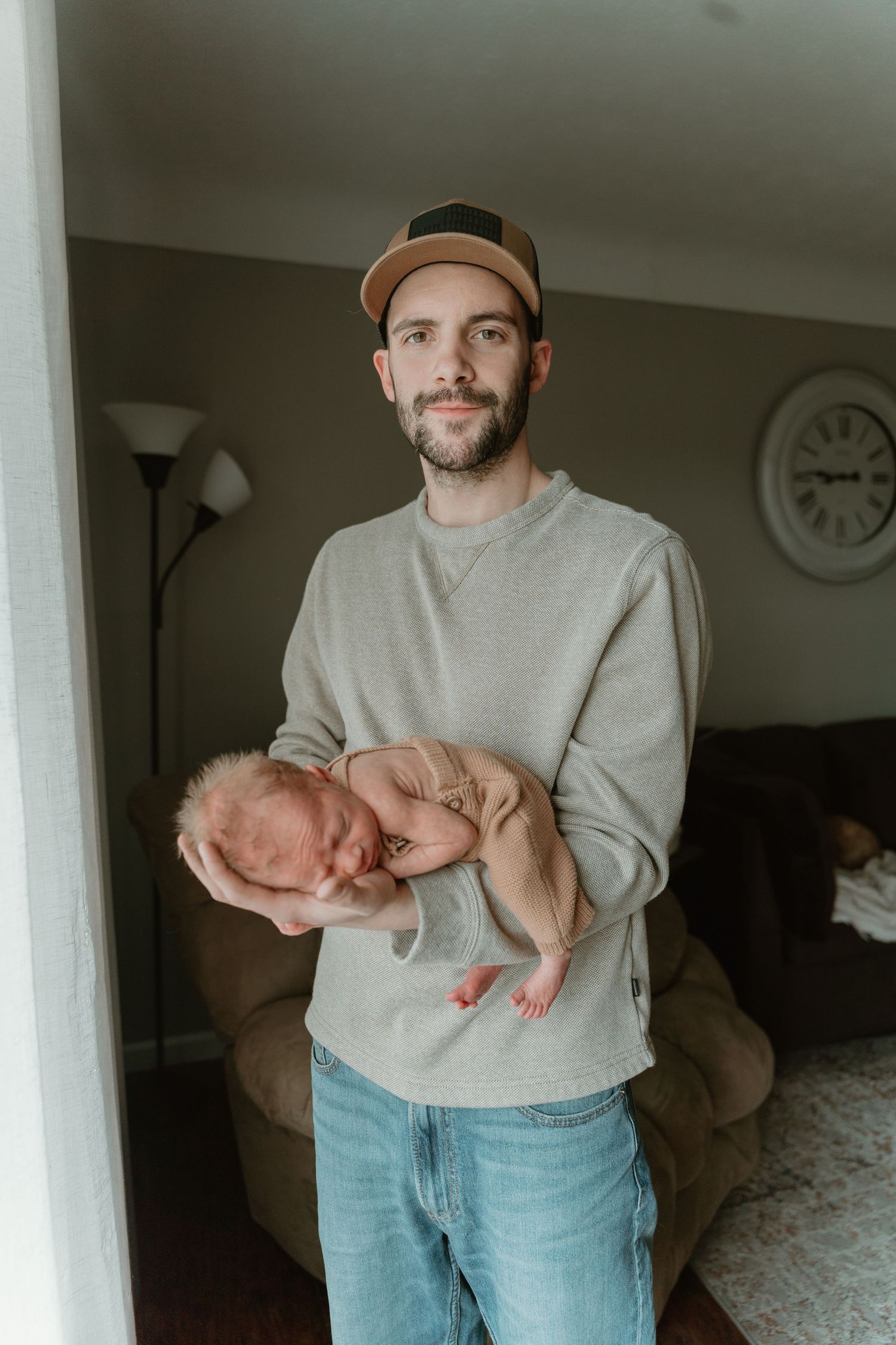 New father in cap and gray sweatshirt proudly holding sleeping newborn baby in home living room.