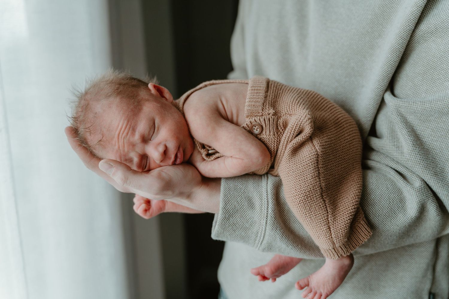 Sleeping newborn baby in tan knit outfit cradled in parent's arms near bright window.