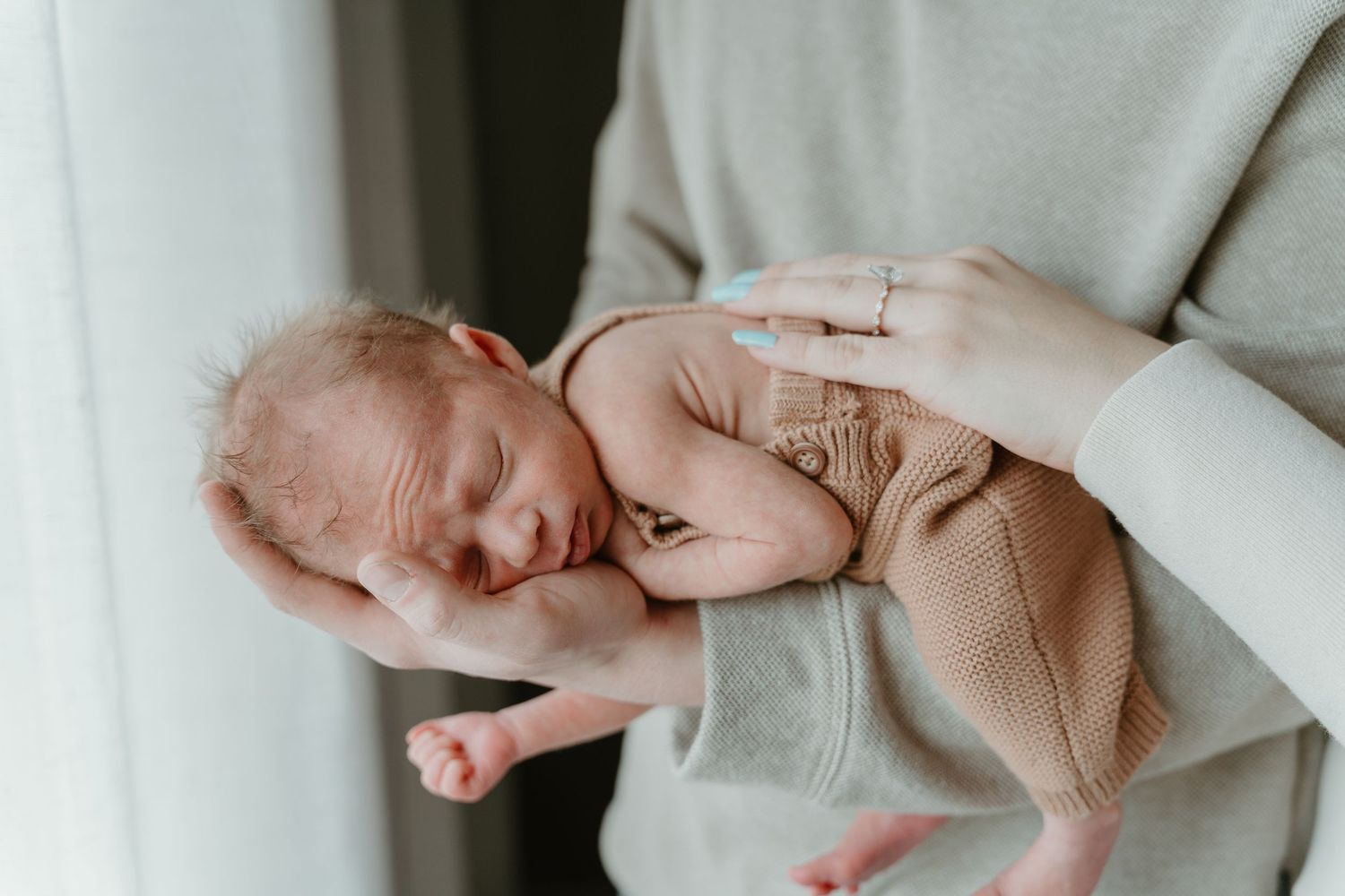 Newborn baby sleeping peacefully in tan knit outfit held gently in parent's hands near window.