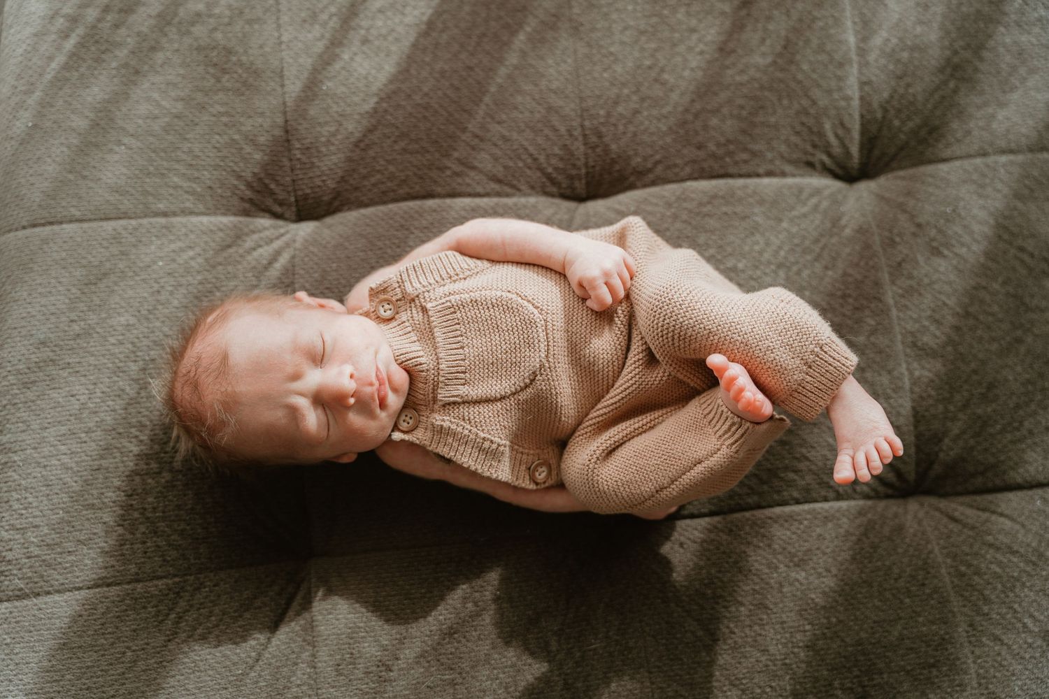 Sleeping newborn baby in a beige knit romper lying on a gray tufted sofa.