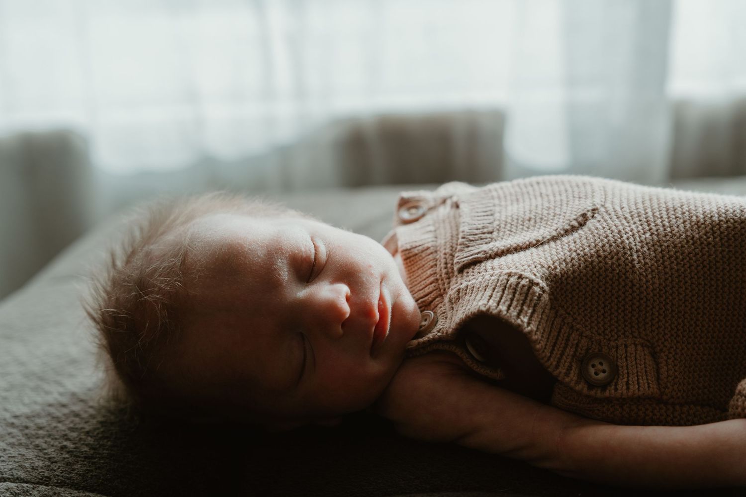 Peaceful newborn baby sleeping in a tan knit outfit on a gray couch near a window.