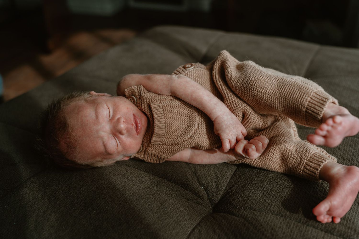 Newborn baby in a brown knit romper lying on a gray couch with legs stretched out.
