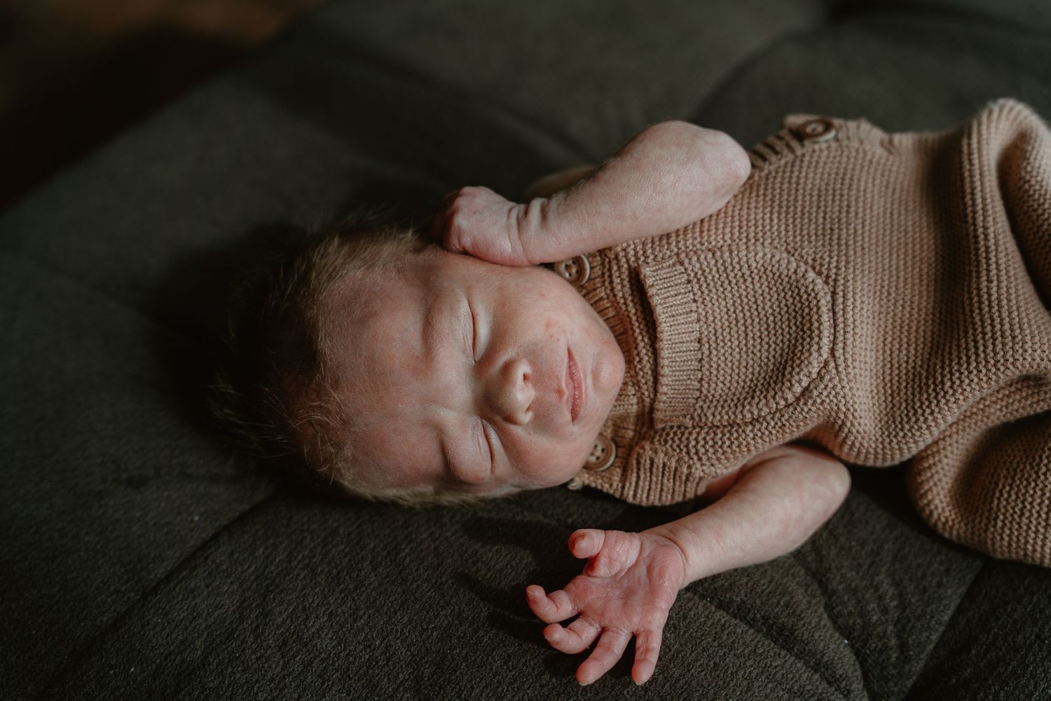Newborn baby in a tan knit romper sleeping with one hand raised beside their head.