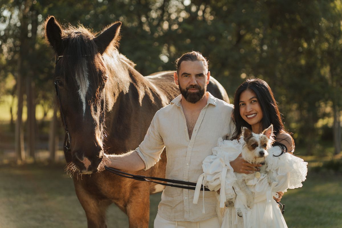 Couple in white outfits posing with a brown horse and small dog in a sunny outdoor setting during a photoshoot with a horse in sydney