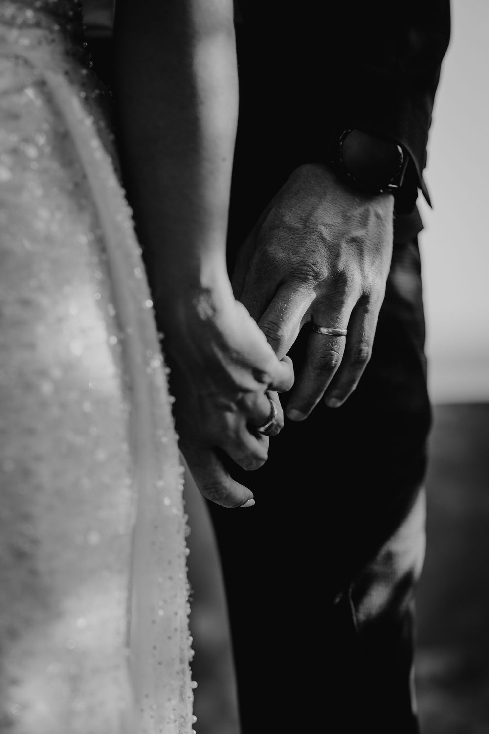 Close-up black and white photo of bride and groom holding hands showing wedding rings and sparkle dress.