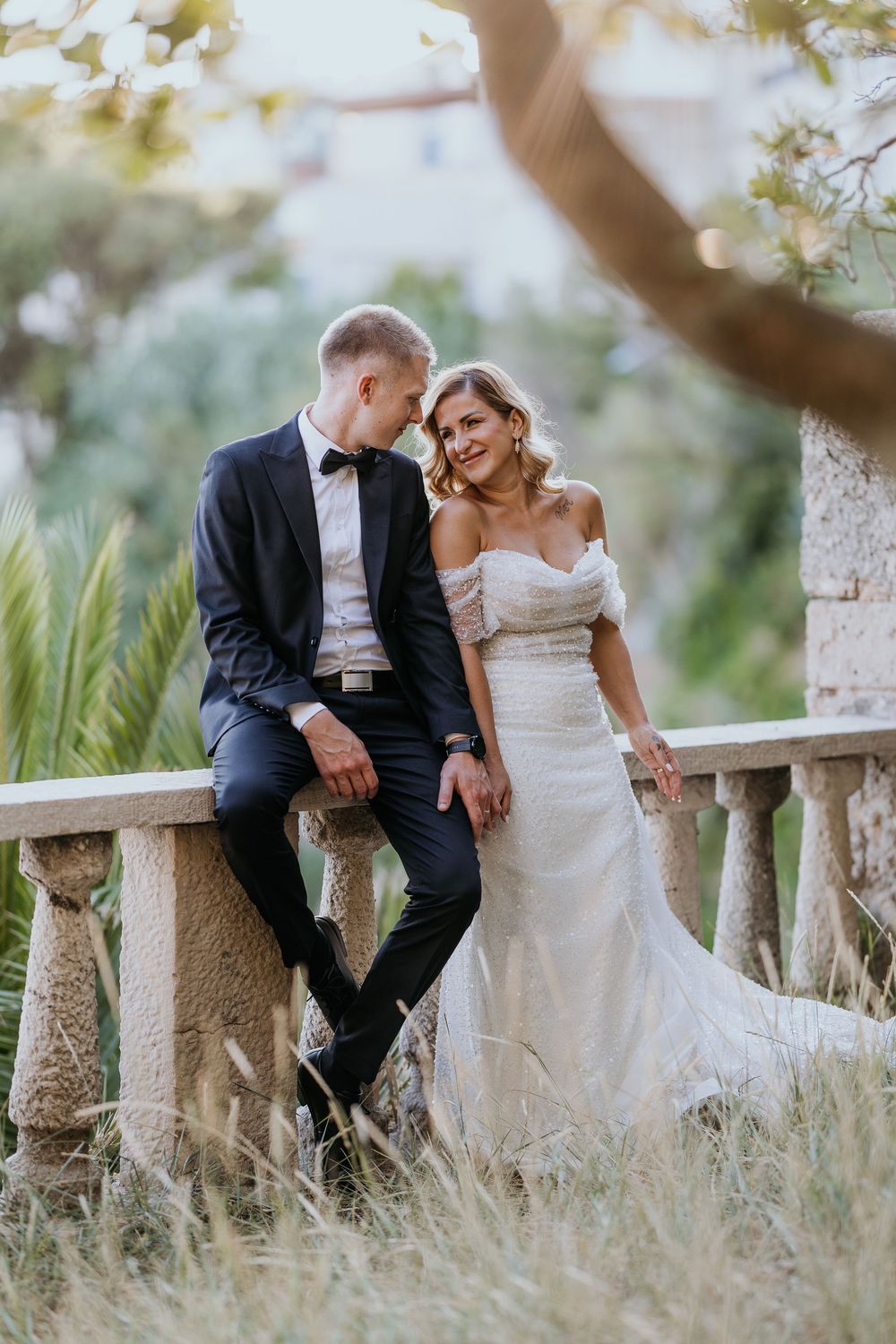 Bride and groom sit together on stone balustrade surrounded by golden hour light.