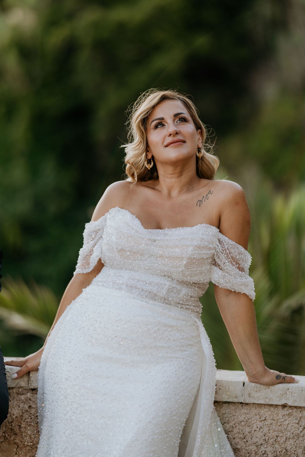 Elegant bride in off-shoulder beaded wedding gown gazes upward in lush garden.