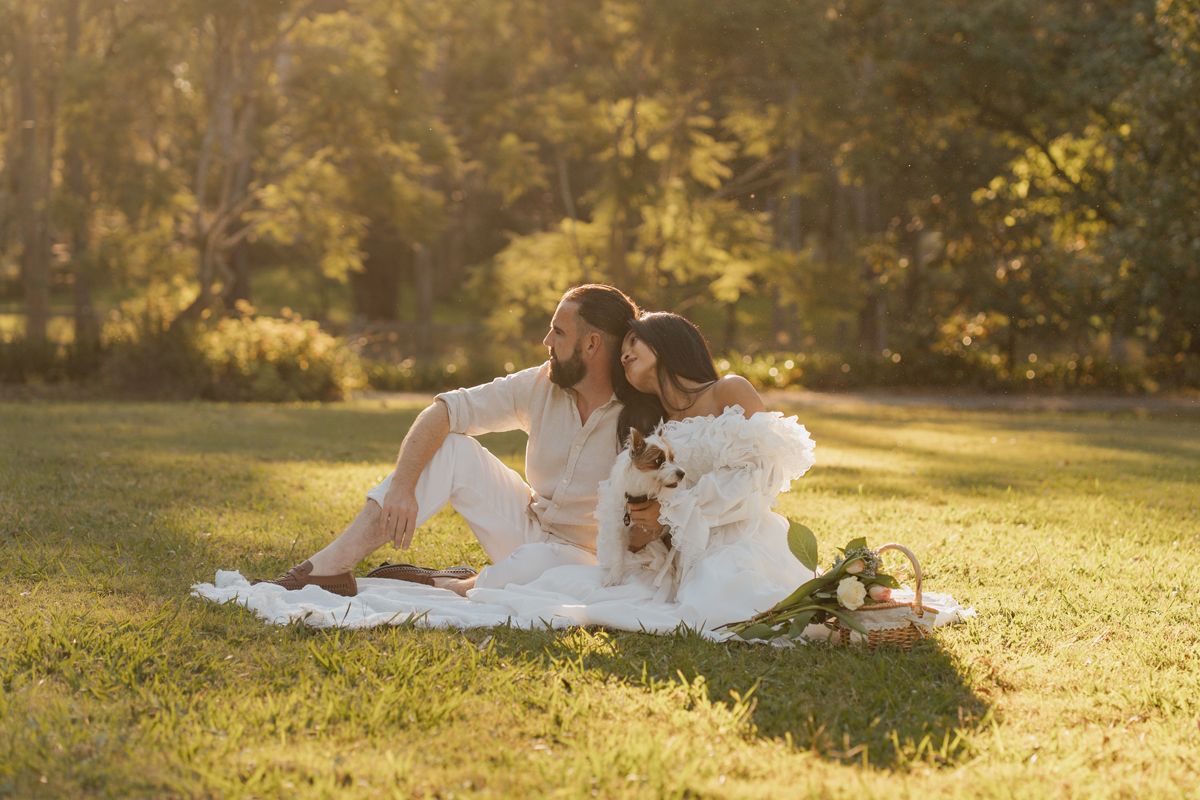 couple in white sit together on a blanket in a sunlit park during a romantic outdoor picnic with their dog on a photoshoot in Rouse hill