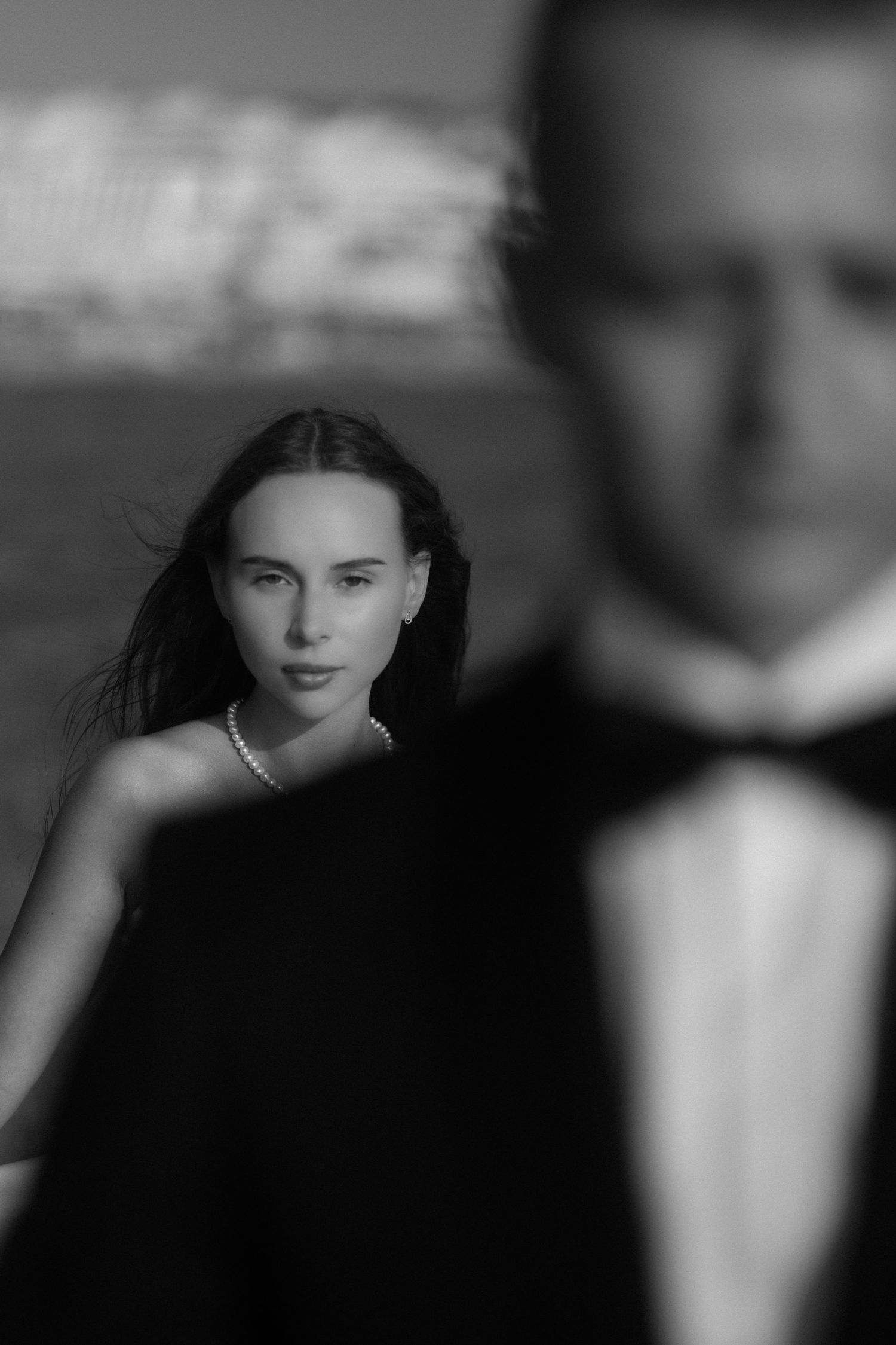 Black and white photo of elegant couple, woman in pearl necklace gazes forward, man in tuxedo blurred in foreground.