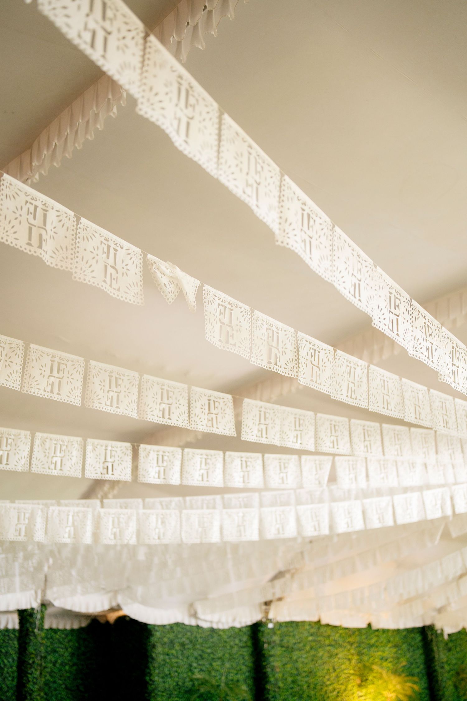 White papel picado banners hanging in diagonal rows against a white ceiling above a green hedge wall.