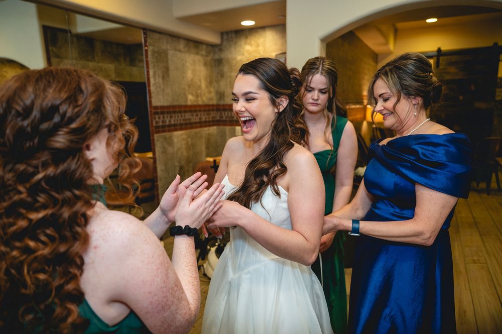 Bride in white gown laughing with bridesmaids in green and blue dresses during wedding celebration.