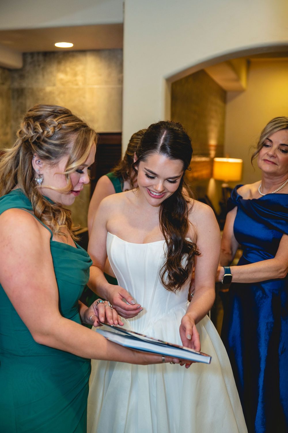 Bride in white strapless gown smiles while reviewing wedding album with bridesmaids in green and blue dresses.