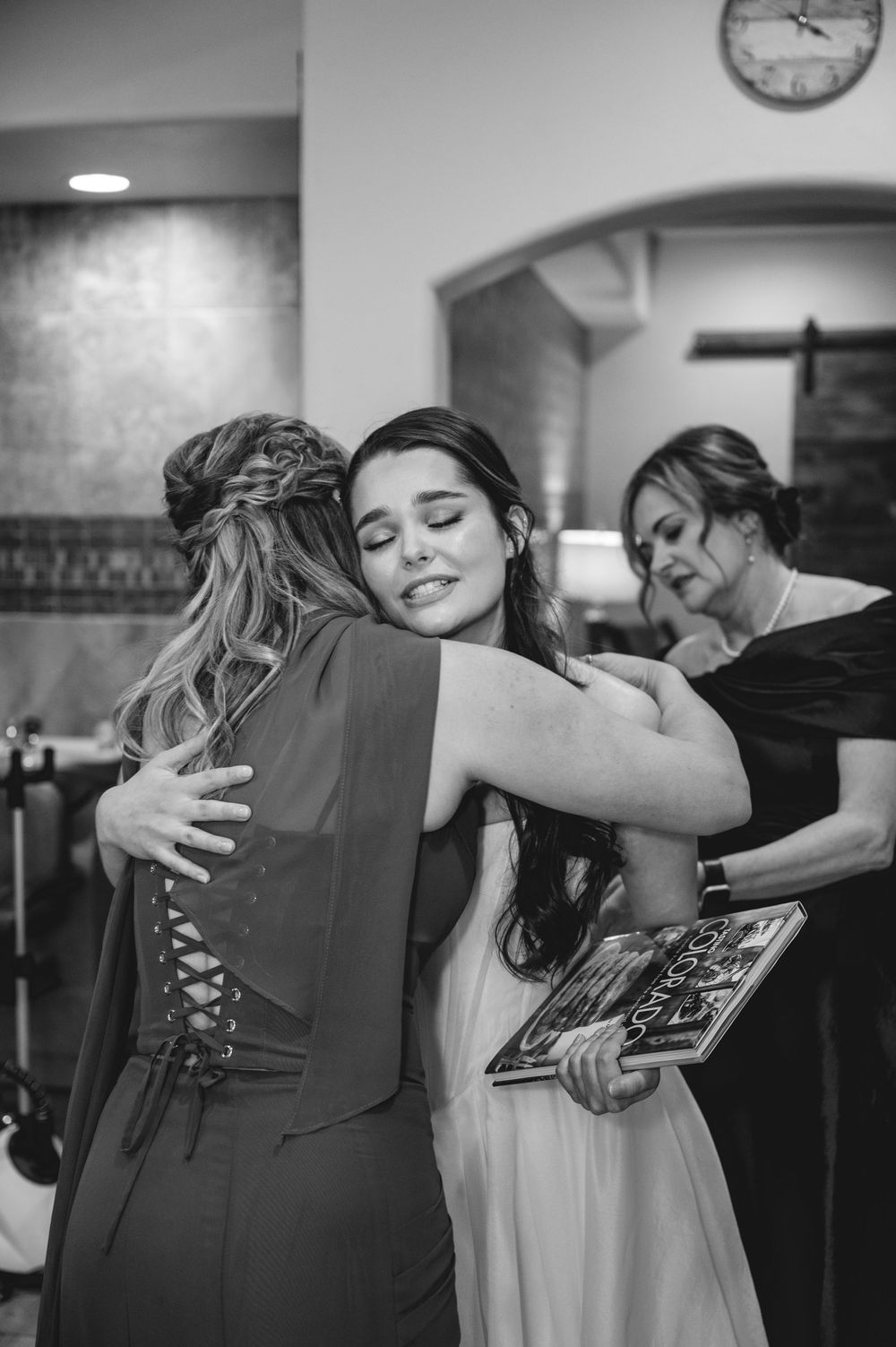 Black and white photo of two women sharing a warm hug at a wedding, with a third woman visible in the background.