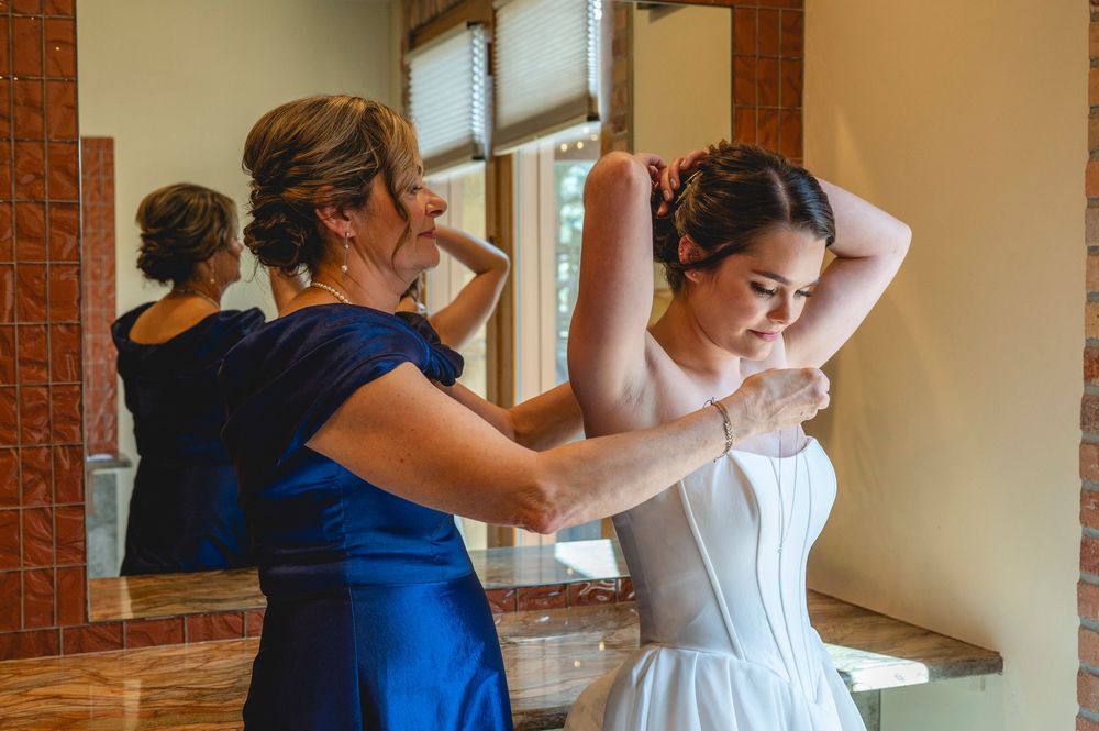 Bride getting help with her white corset wedding dress from a woman in a blue gown in a mirror-lined dressing room.