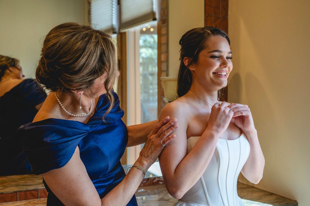 A bride in a white strapless gown smiles as a woman in blue helps her with her dress before the wedding ceremony.