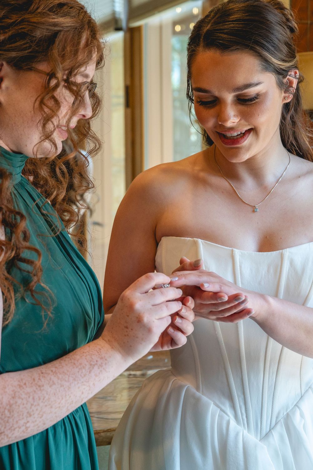 Bride in white strapless gown smiles as bridesmaid in teal dress helps place a ring on her finger.