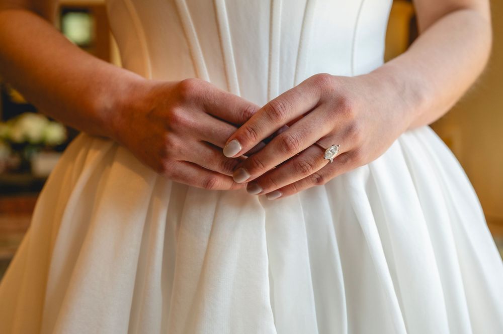 Close-up of bride's hands clasped together wearing a diamond engagement ring on her white wedding dress.