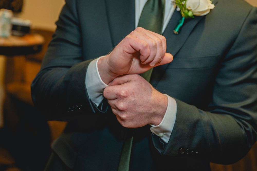 A groom in a dark suit adjusts his cufflinks, wearing a green tie and white boutonniere on his lapel.