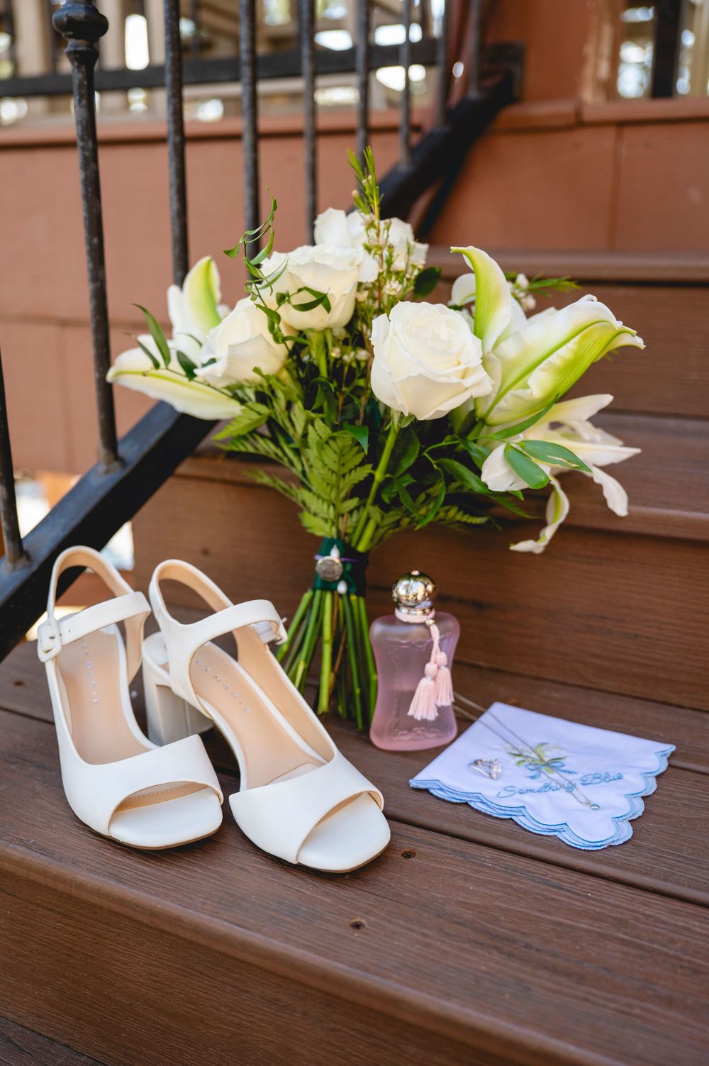 White block heel wedding shoes beside bridal bouquet of roses and lilies, perfume bottle, and handkerchief on wooden steps.