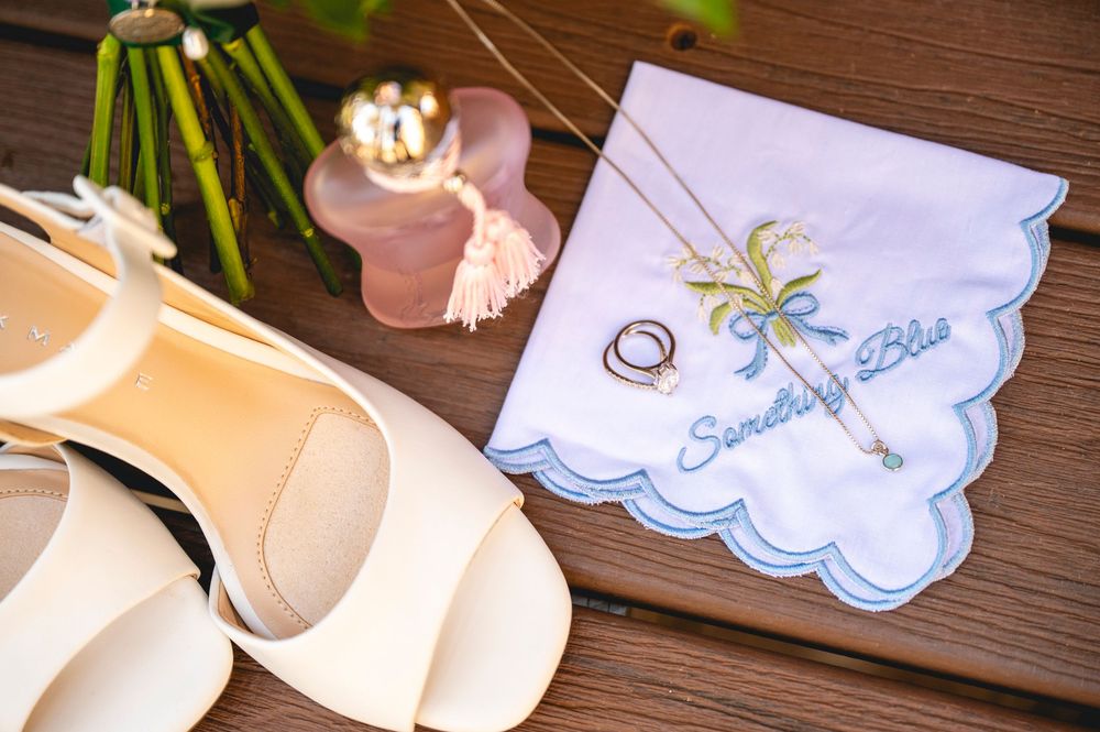 Wedding flat lay with ivory heels, gold rings, palm tree handkerchief, and delicate necklace on wooden surface.