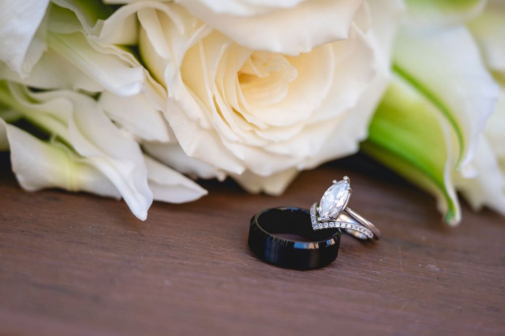 Black tungsten wedding band and diamond engagement ring displayed on wood surface with white roses bouquet backdrop.